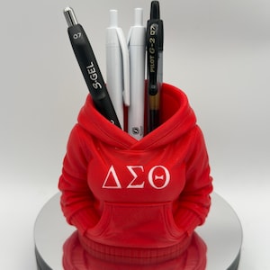 K&ouml;nnte beinhalten: Ein roter Stifthalter in Form eines Hoodies mit den griechischen Buchstaben &Delta;&Sigma;&Theta; in Wei&szlig;. Er enth&auml;lt einen schwarzen S.Gel-Stift, zwei wei&szlig;e Stifte und einen schwarzen Pilot G-2 07-Stift. Der Halter steht auf einer reflektierenden Oberfl&auml;che.