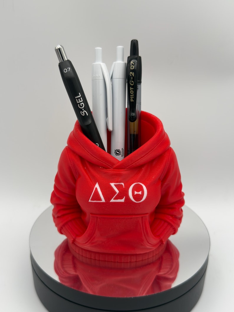 K&ouml;nnte beinhalten: Ein roter Stifthalter in Form eines Hoodies mit den griechischen Buchstaben &Delta;&Sigma;&Theta; in Wei&szlig;. Er enth&auml;lt einen schwarzen S.Gel-Stift, zwei wei&szlig;e Stifte und einen schwarzen Pilot G-2 07-Stift. Der Halter steht auf einer reflektierenden Oberfl&auml;che.