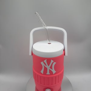 May include: A bright pink insulated beverage cooler featuring a white New York Yankees logo on the front. The cooler has a white lid with a white carrying handle and a clear, curved plastic straw inserted through the center of the lid.