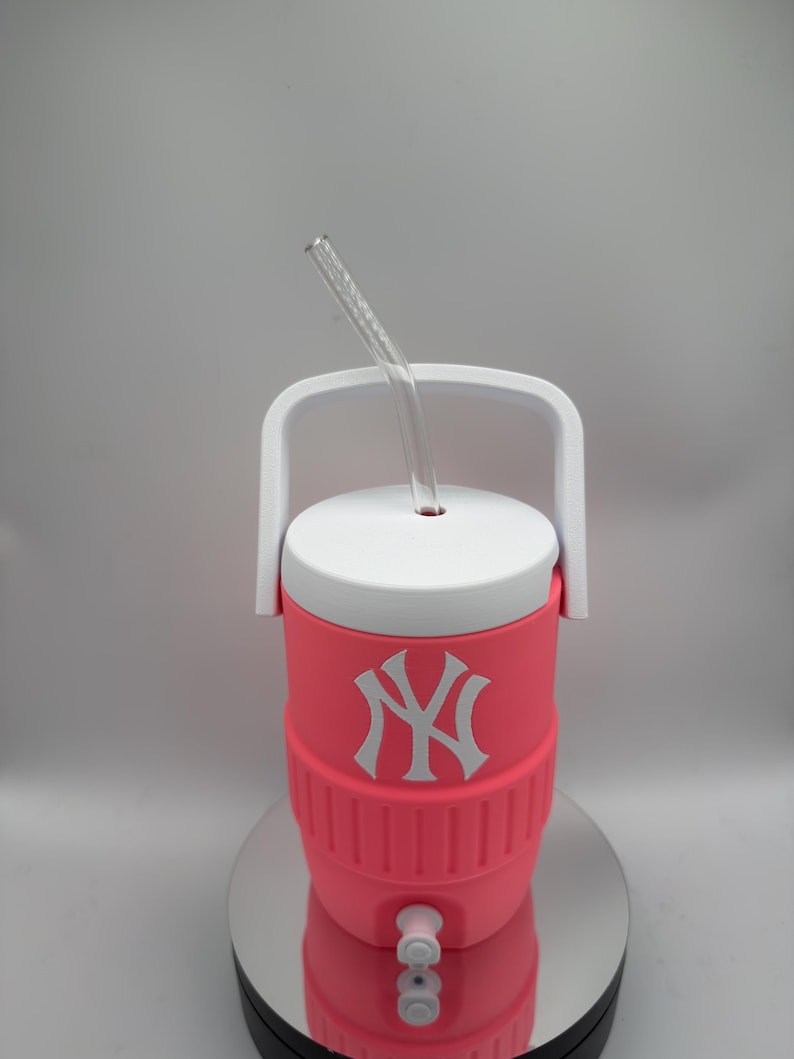 May include: A bright pink insulated beverage cooler featuring a white New York Yankees logo on the front. The cooler has a white lid with a white carrying handle and a clear, curved plastic straw inserted through the center of the lid.