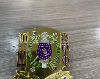 Può includere: Spilla dorata con un emblema viola e verde. La spilla presenta il testo "1911" e "LIFE MEMBER". L'emblema include uno scudo, spade e le lettere greche "ΩΤΦ". Piccoli cristalli trasparenti sono incastonati sui bordi.