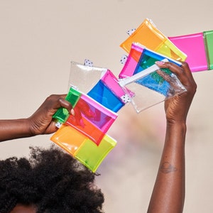 May include: A collection of colorful, transparent pouches in various bright hues, including pink, orange, green, and blue. Each pouch has a zipper closure and a small, patterned tag. The pouches are held up in the air against a neutral background.
