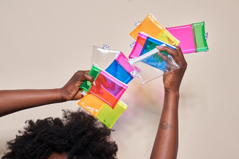 May include: A collection of colorful, transparent pouches in various bright hues, including pink, orange, green, and blue. Each pouch has a zipper closure and a small, patterned tag. The pouches are held up in the air against a neutral background.