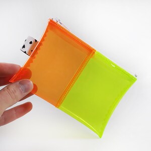 May include: A small, square pouch with a vibrant color scheme. The pouch is divided into two sections, one orange and one neon green, with a white and black patterned tag. The edges are stitched with orange thread.