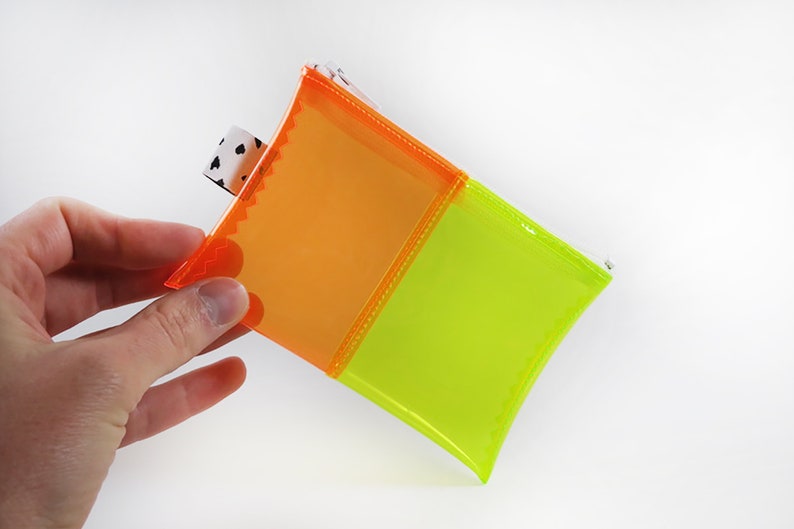 May include: A small, square pouch with a vibrant color scheme. The pouch is divided into two sections, one orange and one neon green, with a white and black patterned tag. The edges are stitched with orange thread.