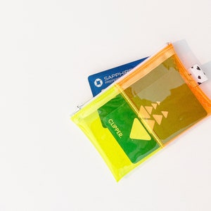 May include: A clear, multi-colored zippered pouch with a blue credit card and a green card that says "CLIPPER." The pouch has a yellow, orange, and clear design with a small black and white heart-patterned tag.