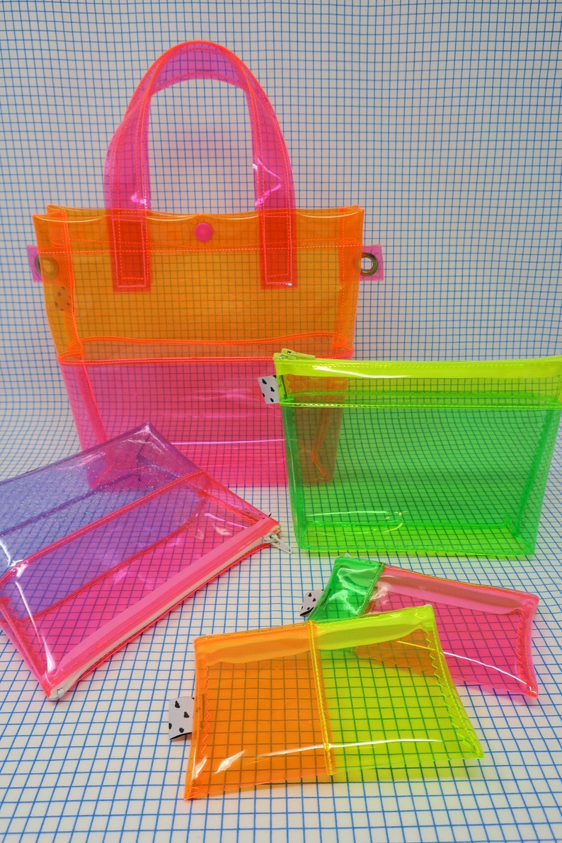 May include: A collection of transparent, neon-colored bags and pouches. The set includes a tote bag, a zippered pouch, and several smaller pouches in shades of pink, orange, and green. The bags have a grid pattern and are made of a clear, flexible material.