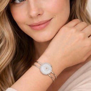 May include: A silver-toned cuff bracelet with a large, round, white stone center and a small gold-colored accent. The bracelet has a double-band design and is worn on a wrist.