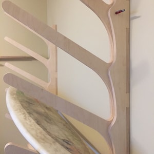 May include: A wall-mounted wooden surfboard rack with a curved design. The rack is made of light-colored wood and has four slots for storing surfboards. A surfboard is resting on the bottom slot.
