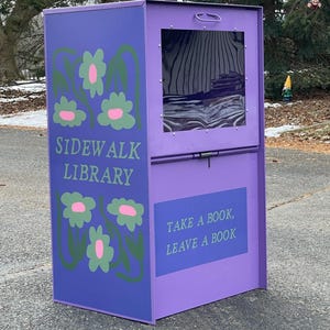 Puede incluir: Una biblioteca de acera morada con ventana y puerta. El lateral de la biblioteca tiene diseños de flores verdes y rosas y las palabras "SIDEWALK LIBRARY". La parte inferior de la biblioteca dice "TAKE A BOOK, LEAVE A BOOK".