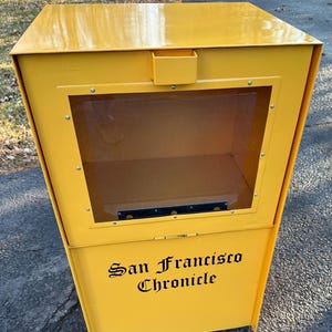 May include: A yellow metal newspaper box with a clear plastic window and a slot on the top. The box has the words "San Francisco Chronicle" printed on the front in black.