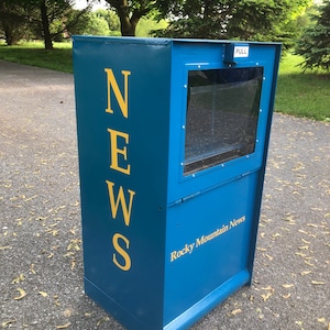 May include: A blue metal newspaper box with a clear plastic window and a yellow "NEWS" sign. The box has a "PULL" sign and the text "Rocky Mountain News" on the front.