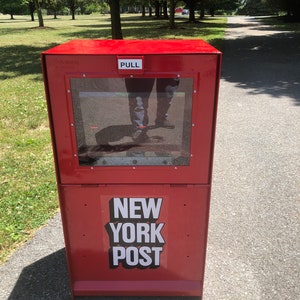 May include: A red metal newspaper box with a glass window and a sign that says "NEW YORK POST" on the front.