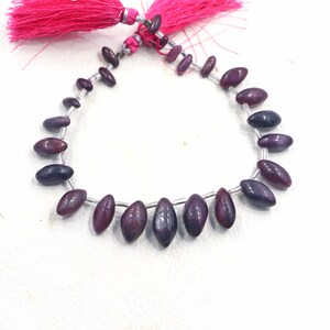 May include: A strand of oval-shaped, dark purple beads with a metallic silver string. The beads are a mix of deep purple and burgundy hues. A pink tassel is attached to the top of the strand.