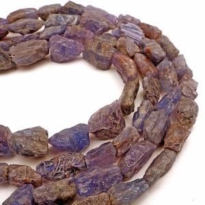 May include: Strands of rough-cut tanzanite gemstones in shades of purple, blue, and brown. The raw, irregular shapes of the stones are strung together, creating a natural and earthy aesthetic. The gemstones are displayed against a white background.