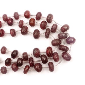 May include: A strand of faceted, round, deep red ruby beads strung on a thin, clear filament. The beads exhibit a translucent quality with natural variations in color and texture, arranged in a repeating pattern along the length of the string.