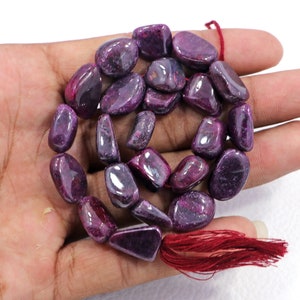 May include: A strand of purple ruby gemstone beads, each bead is a tumbled, freeform shape. The beads are strung on a cord with a red tassel at the end.