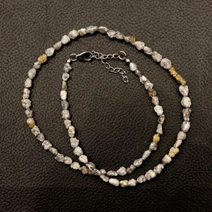 Può includere: Una collana composta da numerose piccole pietre grezze e irregolari nei toni del grigio, bianco e giallo pallido. Il filo presenta una chiusura a moschettone in metallo argentato e una catenella di estensione regolabile per la lunghezza.