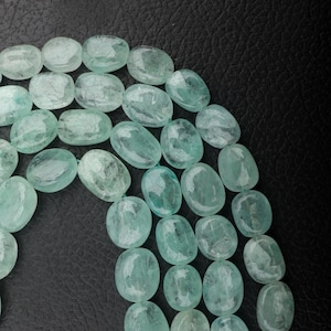 Könnte beinhalten: Nahaufnahme eines Strangs ovaler Aquamarinperlen. Die Perlen haben eine hellblau-grüne Farbe und sind aufgefädelt. Der Hintergrund ist eine dunkle, strukturierte Oberfläche, die einen Kontrast zu den Perlen bildet.