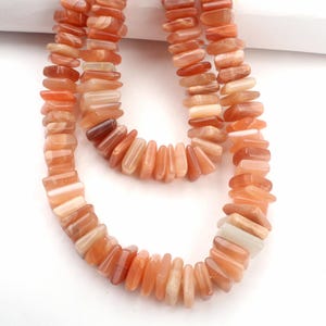 Puede incluir: Un collar hecho de cuentas rectangulares de color naranja y beige. Las cuentas están ensartadas para formar un collar. El collar se muestra sobre un fondo blanco.
