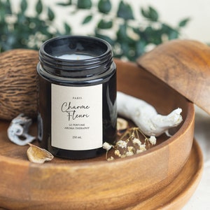 May include: A black glass candle jar with a white label that reads "Paris Charme Fleuri Le Perfume Aroma Therapy 250 ml". The candle is sitting in a wooden bowl with a white piece of driftwood and white flowers.