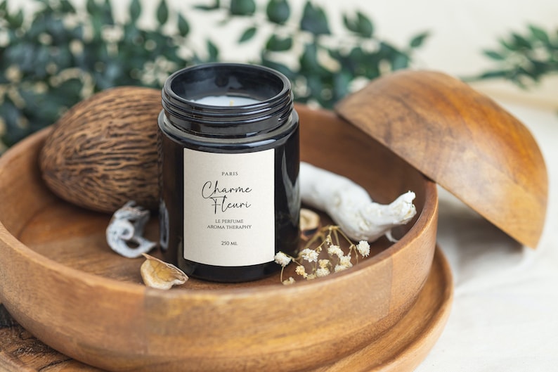 May include: A black glass candle jar with a white label that reads "Paris Charme Fleuri Le Perfume Aroma Therapy 250 ml". The candle is sitting in a wooden bowl with a white piece of driftwood and white flowers.