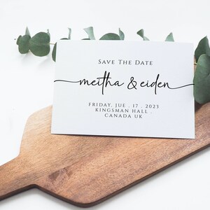 May include: A white save the date card with black text. The text reads "Save the Date" followed by "meitha & ciden" in a cursive font. The date, location, and city are listed below the names. The card is sitting on a wooden cutting board with a sprig of eucalyptus leaves.