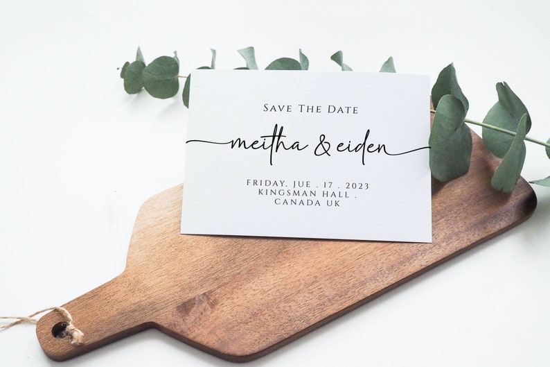 May include: A white save the date card with black text. The text reads "Save the Date" followed by "meitha & ciden" in a cursive font. The date, location, and city are listed below the names. The card is sitting on a wooden cutting board with a sprig of eucalyptus leaves.