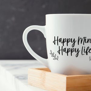 May include: White ceramic mug with black text that reads "Happy Mind Happy Life" and small black leaf designs on either side of the text. The mug is sitting on a wooden coaster.