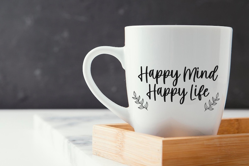 May include: White ceramic mug with black text that reads "Happy Mind Happy Life" and small black leaf designs on either side of the text. The mug is sitting on a wooden coaster.