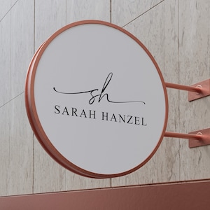 Op de afbeelding: Een rond, wit bord met een koperen frame en een zwart, cursief logo dat "sh" zegt en daaronder "SARAH HANZEL". Het bord is aan een muur bevestigd met koperen beugels.