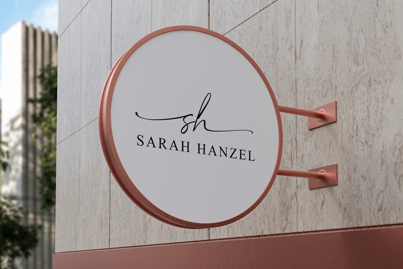 Op de afbeelding: Een rond, wit bord met een koperen frame en een zwart, cursief logo dat "sh" zegt en daaronder "SARAH HANZEL". Het bord is aan een muur bevestigd met koperen beugels.