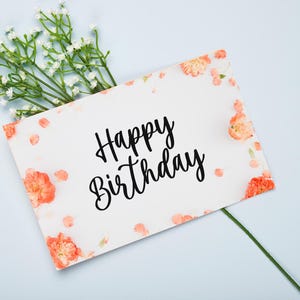 May include: A white card with the words "Happy Birthday" written in black cursive script. The card is decorated with small pink flowers and white petals.