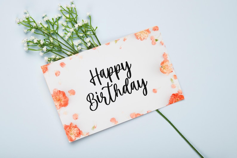 May include: A white card with the words "Happy Birthday" written in black cursive script. The card is decorated with small pink flowers and white petals.