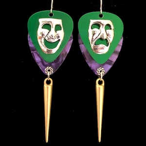 May include: A pair of dangle earrings featuring green and purple guitar picks with silver comedy and tragedy masks. The earrings have gold spikes hanging from the bottom.