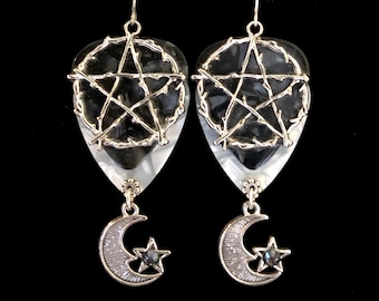 May include: A pair of drop earrings featuring a black and white teardrop-shaped pendant with a silver pentagram design. Each earring has a silver crescent moon and star charm with a dark stone accent. The earrings hang from silver hooks.