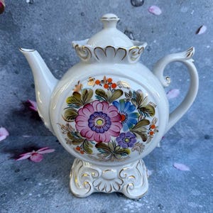 May include: A white ceramic teapot with a floral design. The teapot features a round body, a curved spout, and a handle. The floral design includes pink, blue, and purple flowers with green leaves. The teapot sits on a decorative base with gold accents.