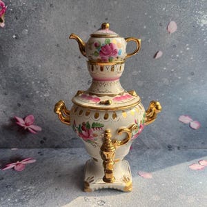 May include: A white porcelain samovar featuring a floral pattern with pink roses and green leaves, accented by gold trim on the handles, spout, and base. A small matching teapot sits securely on top of the samovar, completing the set.