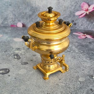 May include: A golden, ornate samovar with black accents. The tea kettle features a round body, a lid with a small handle, and a spigot. The samovar is displayed on a gray surface with pink flower petals scattered around it.