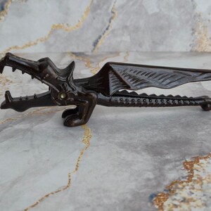 May include: A dark brown dragon-shaped nutcracker with detailed wings and a hinged jaw. The nutcracker is made of metal and has a decorative design, perfect for cracking nuts. The dragon's mouth is open, revealing teeth.