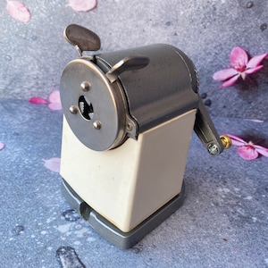 May include: A vintage silver pencil sharpener with a white plastic base and a metal crank. The sharpener has a round blade and a small hole for sharpening pencils.
