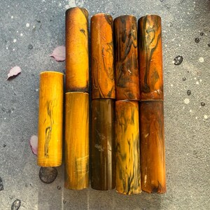May include: A collection of eight cylindrical objects arranged in two rows. The pieces feature a variety of warm tones including golden yellow, amber, and deep brown, with marbled or swirled patterns throughout the smooth, polished surfaces.