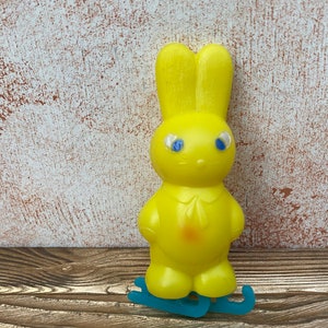 May include: A yellow plastic toy bunny wearing blue and white eyes, standing on blue plastic skates. The bunny has a white collar and a pink spot on its chest.