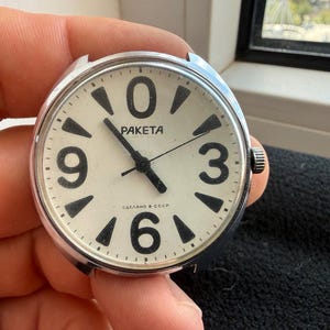 Peut inclure: Un cadran de montre rond argenté avec un fond crème, présentant de grands chiffres noirs aux positions 0, 3, 6 et 9. Le cadran affiche le texte PAKETA et СДЕЛАНО В СССР, avec des aiguilles noires pour les heures, les minutes et les secondes.
