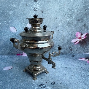 May include: A silver-colored samovar with a spigot, handles, and a lid with a decorative top. The tea kettle sits on a square base. The background is a mottled blue-gray with pink flower petals.