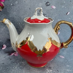 May include: A white teapot with a red and gold design. The teapot has a gold handle and spout. The teapot is sitting on a gray surface.