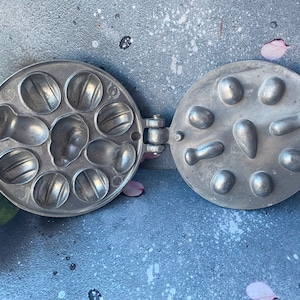 May include: A vintage metal waffle iron with two hinged sections. One side has eight round molds for making small waffles, the other side has eight molds for making different shapes.