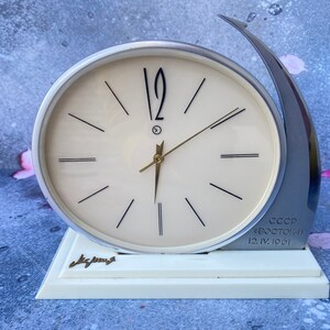 Pode incluir: Um relógio de mesa branco e prateado com um design curvo. O mostrador do relógio tem ponteiros de hora e minuto pretos e marcas de números romanos pretos. O mostrador do relógio também tem o texto "CCCP "СПОСТОИЧ" 12.IV.1961". O relógio está sobre uma base branca com a palavra "Чacous" escrita nela.