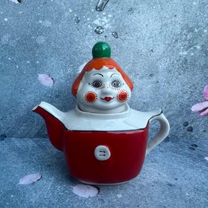 May include: A whimsical red and white ceramic teapot with a clown face design. The teapot features a red body, white collar, and a clown face lid with orange hair, a green knob, and painted facial details. A unique kitchenware item.