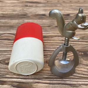 May include: A silver squirrel nutcracker with a red and white plastic container. The nutcracker is made of metal and has a squirrel design on the top. The container is a small, cylindrical container with a red top and a white bottom.